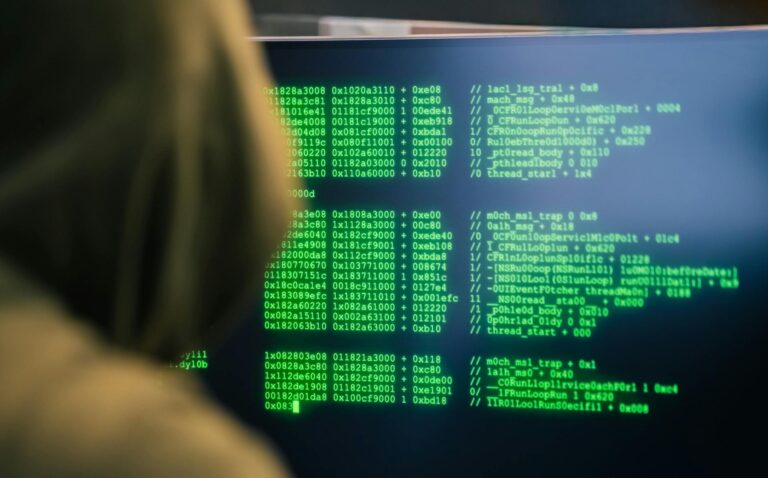 Osoba w zielonej bluzie z kapturem siedząca przed monitorem komputera wyświetlającym zielony kod asemblerowy – symbol hakera i cyberataku